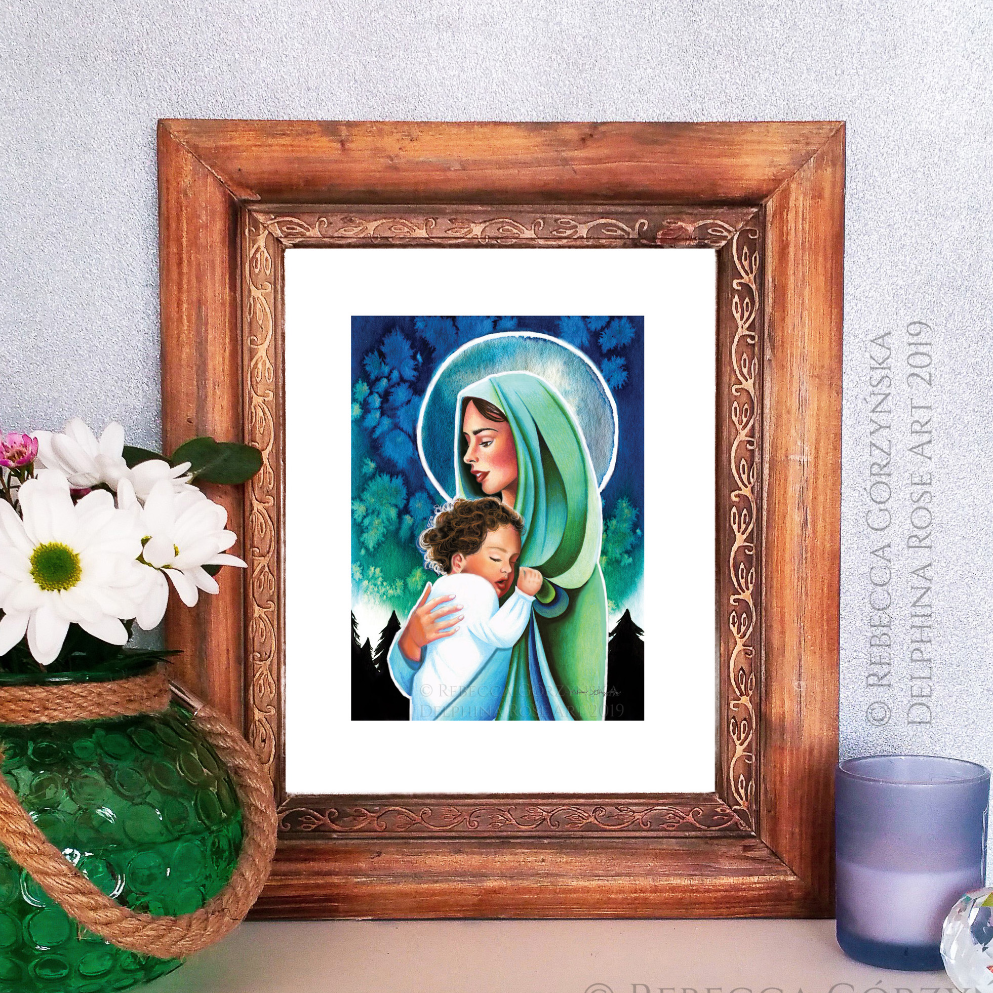 Mary wearing green veil holding sleeping toddler baby Jesus in front of a green-blue watercolor sky and black pine trees in brown wooden frame with vase of flowers