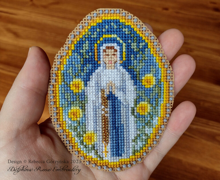 Hand holding an egg-shaped cross stitch ornament stitched on perforated paper. Mary in a whte dress, hands folded in prayer, stands against a blue background and is surrounded by yellow roses. On her arm is a rosary of gold beads, Catholic cross stitch design by Delphina Rose Embroidery